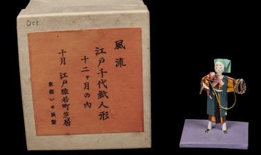 Paper figure of a man holding a monkey on a lead next to a brown and orange box with Japanese characters on the front. 