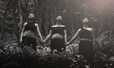 Black and white photograph showing three women standing holding hands in a forest setting