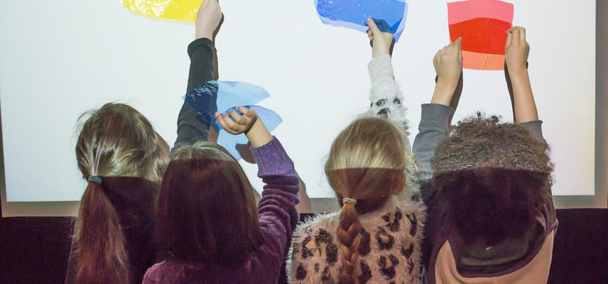 Four children stand in front of a white screen holding up yellow, blue and red translucent plastic sheets to make shadows.