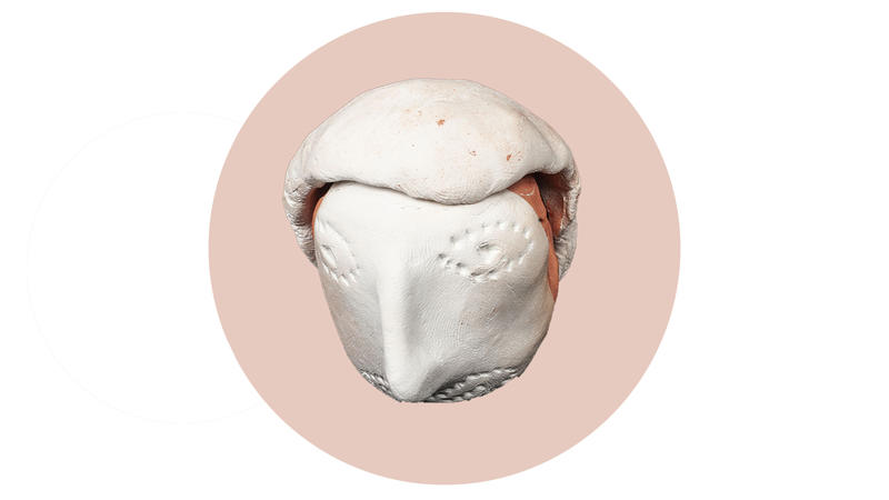 A head moulded out of white clay with a long thin nose pinched into shape and two eyes and a mouth marked with a series of dots. A thin, second piece of clay has been wrapped over the top of the head.