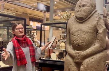 A woman gestures towards a weathered stone statue of human figure carved with its hands in its lap