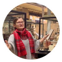 A woman stands among glass display cases with her arms open, caught in the moment of talking about museum objects on display